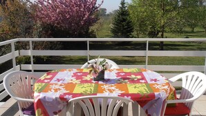 A round table with a colorful tablecloth, surrounded by white chairs, on a balcony with a view of trees and a grassy field.