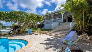 Ein Poolbereich mit Steinboden, eine überdachte Außenbereich-Sitzgelegenheit mit Stühlen und eine Treppe, die zu einem Haus führt, umgeben von Grünflächen.