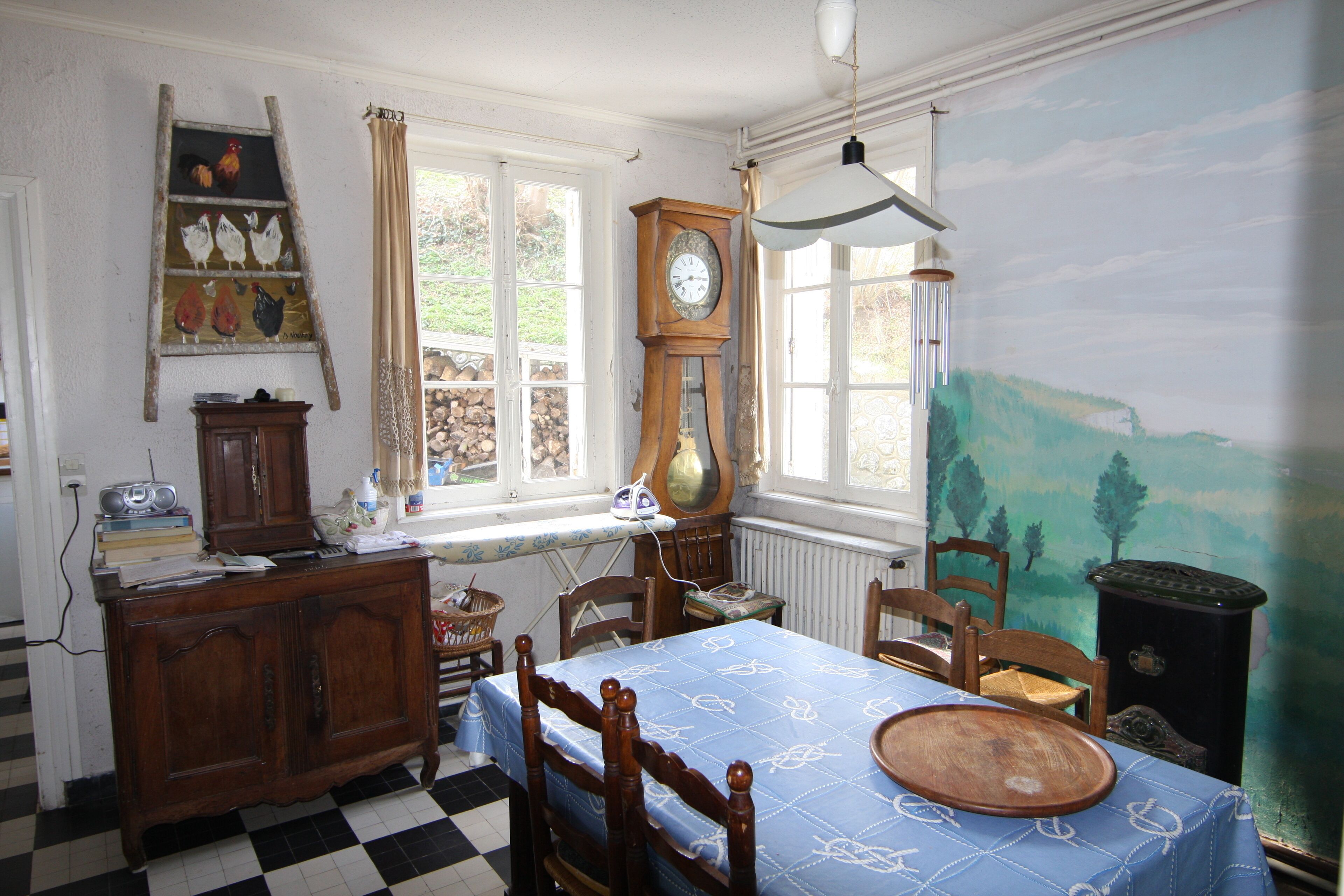 Huoneessa on ruutukuviollinen lattia, sinisellä pöytäliinalla peitetty puupöytä, puinen kaappi, kello ja seinällä suuri seinämaalaus.