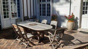 Un bâtiment de style européen traditionnel, avec une façade blanche, une grande table en bois entourée de chaises et un pot de fleurs orné de fleurs rouges.