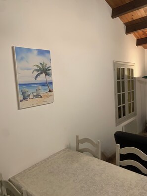 Une pièce avec un mur blanc, un tableau représentant une scène de plage, un meuble blanc et une table avec deux chaises.