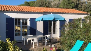 A white building with a red-tiled roof, a table with chairs, a barbecue, and blue lounge chairs.