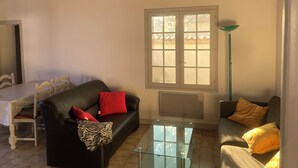 A living room with a black leather sofa, a glass coffee table, and a grey sofa with yellow cushions. There is a window with frosted glass and a green floor lamp.