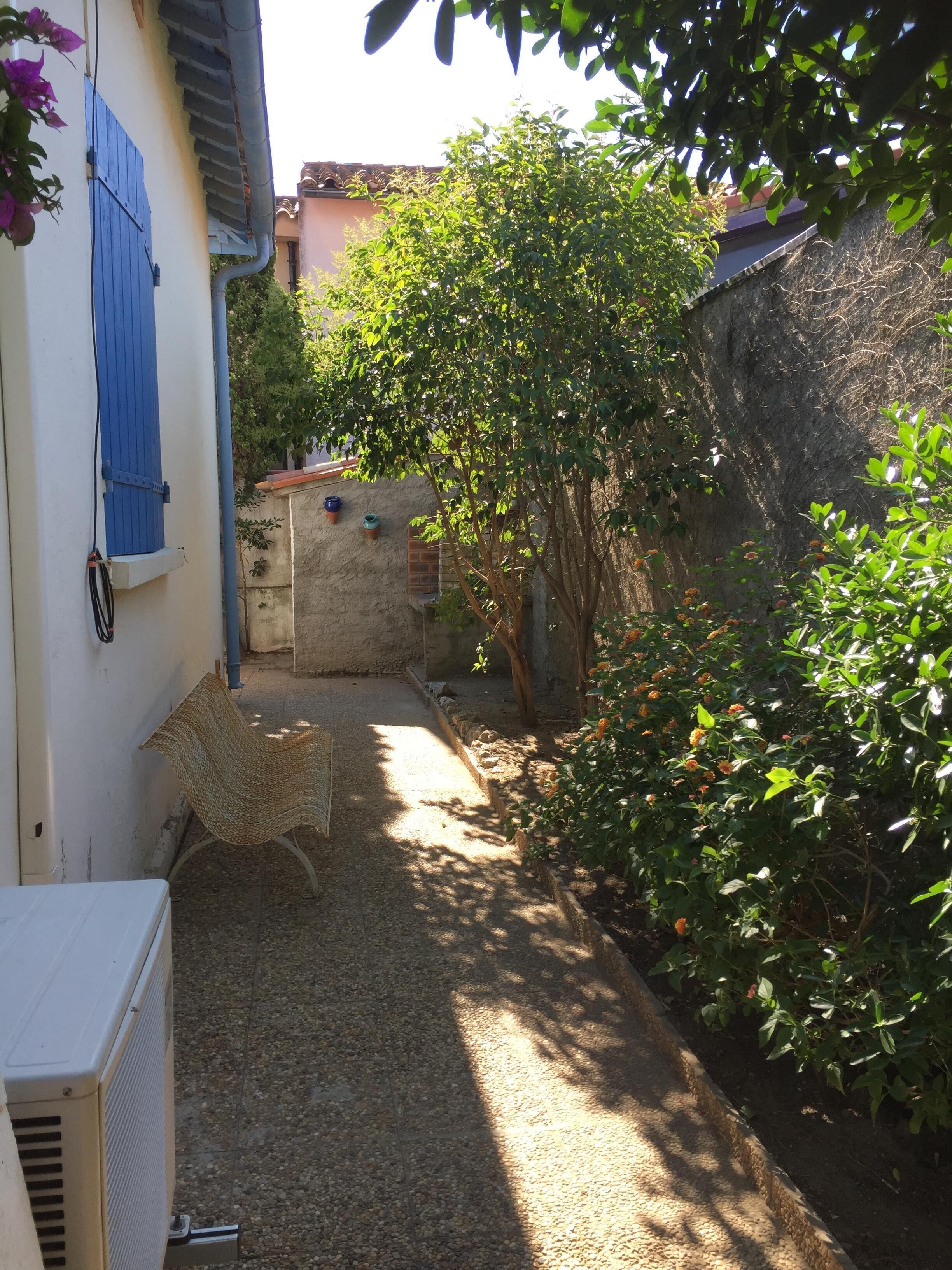A narrow courtyard with a gravel path, a wicker chair, and a white air conditioning unit.