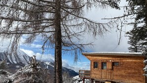 A wooden cabin with a balcony, surrounded by snow-covered trees and mountains in the background.