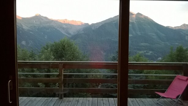 A balcony with a wooden railing and a pink chair, overlooking a mountainous landscape with trees and a clear sky.
