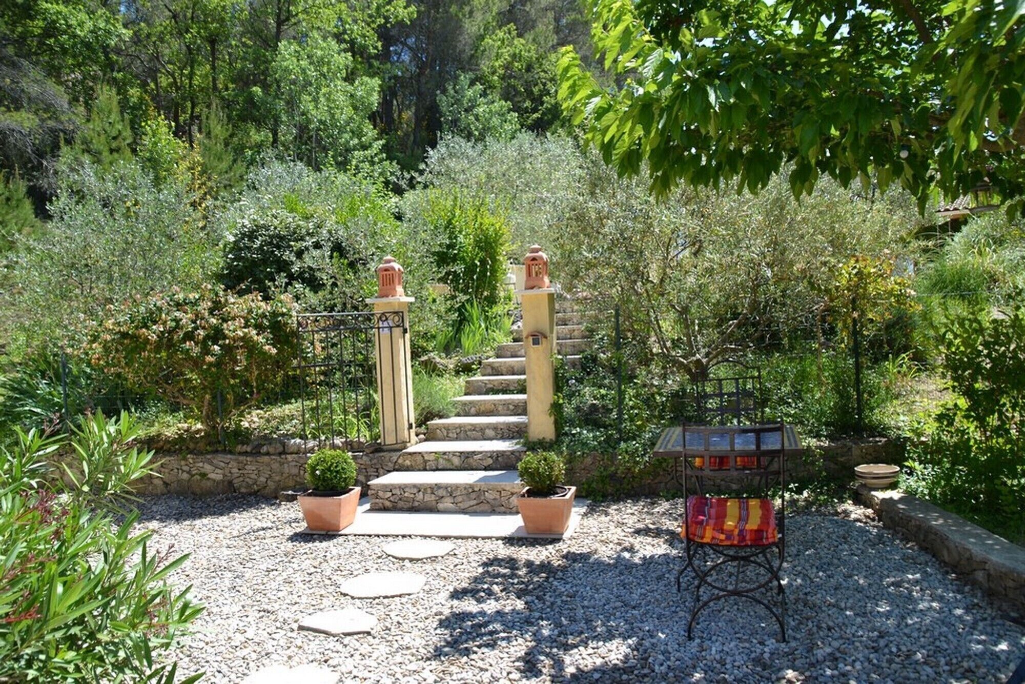 A garden with a stone staircase leading to a gate, surrounded by potted plants and a small table with a colorful cushion.