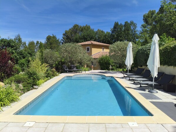 Ein rechteckiger Pool mit gefliestem Rand, umgeben von Liegestühlen und Sonnenschirmen, mit einem Gebäude im Hintergrund.