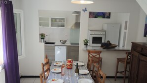 Un coin repas avec une table dressée pour quatre personnes, une cuisine en arrière-plan et une fenêtre ornée de rideaux violets.