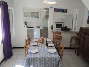 Un comedor con una mesa dispuesta para cuatro, una cocina al fondo y un ventanal con cortinas moradas.