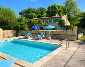 Una piscina con sillas de descanso y sombrillas, rodeada de un muro de piedra y árboles.