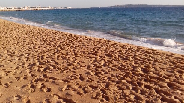 A sandy beach with footprints leading to the water, a clear sky, and a distant shoreline with buildings. -  Beautiful Cannes Apartment (Cannes)