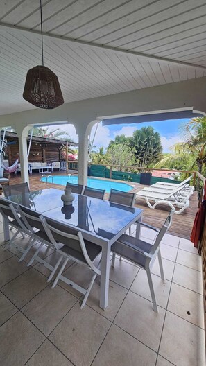 An outdoor patio with a glass table and chairs, a hanging light fixture, and a view of a swimming pool and greenery. - Comfortable villa, swimming pool, tropical garden, near beach st Anne, 8 pers (Sainte Anne)