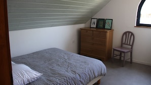 Ein Schlafzimmer mit schrägem Dach, einem Bett mit gestreifter Bettdecke, einem hölzernen Schminktisch, einem Stuhl und einem Fenster mit Aussicht.