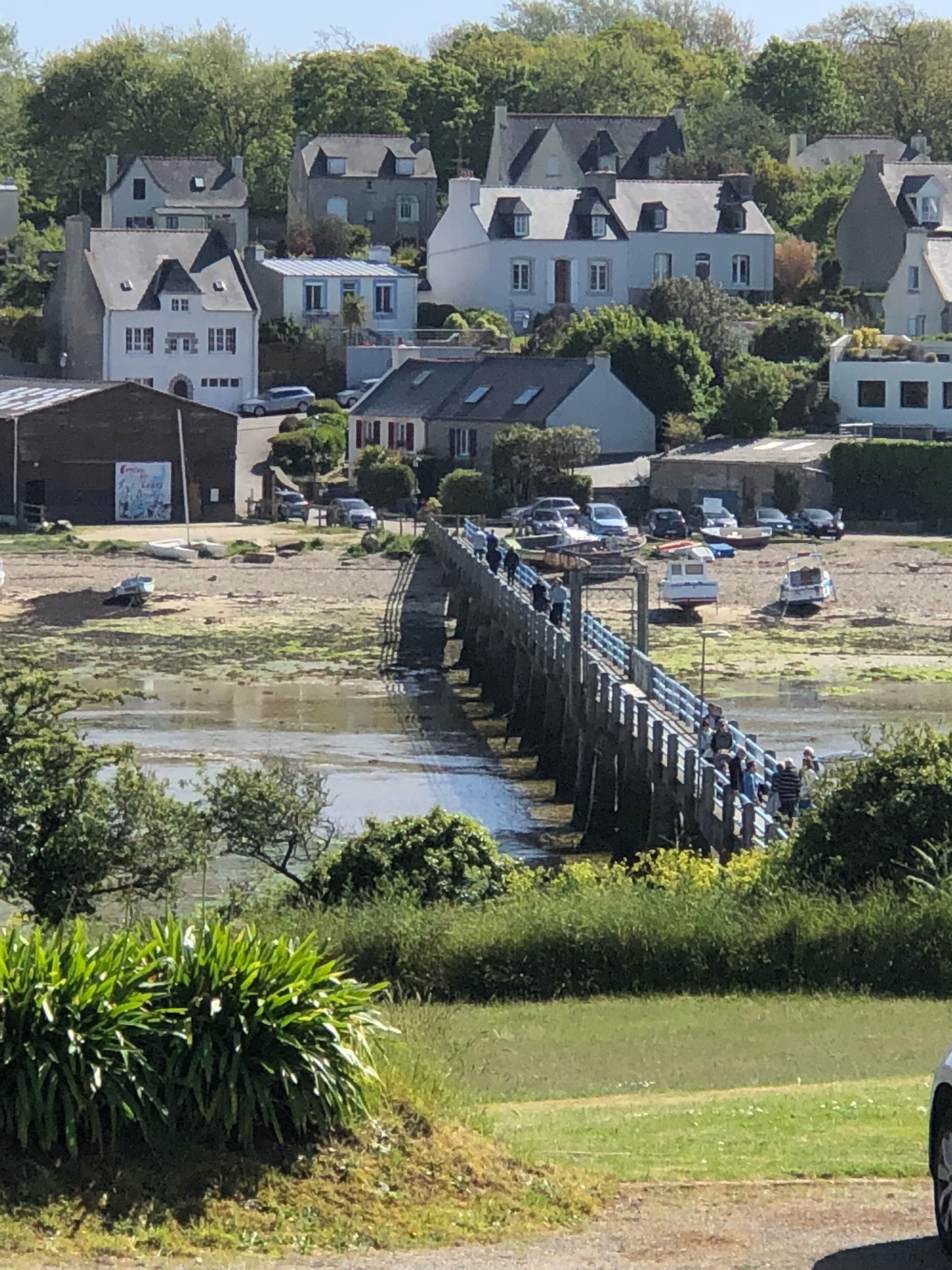 A coastal town with a stone bridge leading to a sandy beach, surrounded by residential houses and parked cars.