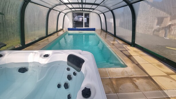 Een overdekt zwembad met een jacuzzi, omgeven door glazen wanden en een betegelde vloer.