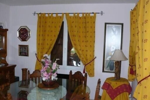 A dining area with a glass table, wooden chairs, yellow curtains, and a lamp on a side table.