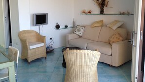 Un soggiorno con un divano color beige, poltrone in vimini, una TV a schermo piatto e oggetti decorativi sugli scaffali.