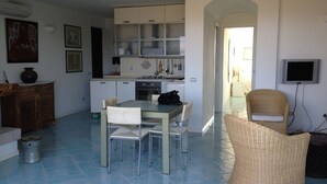 Una cucina moderna con mobili bianchi, un tavolo da pranzo con sedie, una sedia in vimini e un televisore montato a parete.