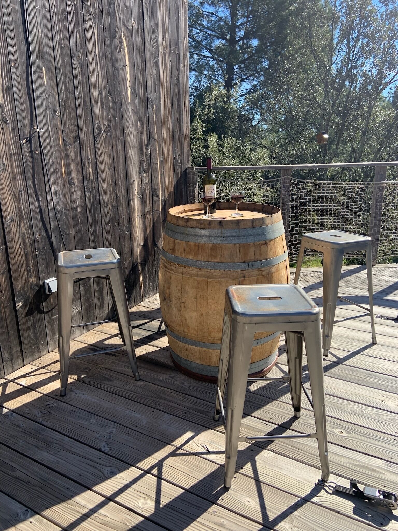 Une terrasse en bois avec un grand tonneau, deux tabourets en métal et une table en métal.