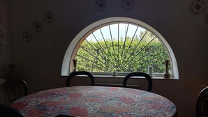 Ein Essbereich mit einem runden Tisch, der mit einer gemusterten Tischdecke bedeckt ist, zwei Stühle, einem Fenster mit Blick auf Grünflächen und einer Vase auf dem Fensterbrett.