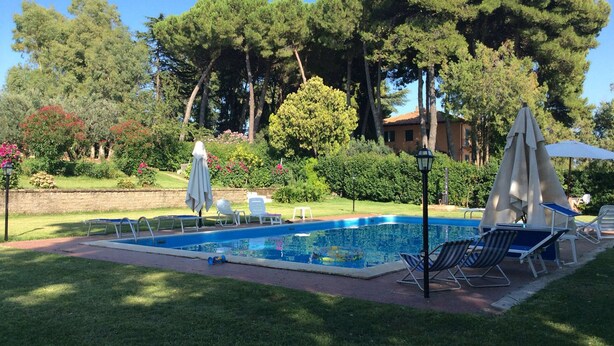 Ein rechteckiger Swimmingpool mit einer gefliesten Liegefläche, umgeben von Gras und Bäumen. Liegestühle und Sonnenschirme befinden sich rund um den Pool.