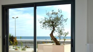 Un salon moderne avec un canapé et un fauteuil blanc original, une grande porte-fenêtre coulissante donnant sur un balcon avec vue sur la mer et un arbre.