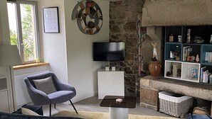 A modern living room with a grey sofa, a small armchair, a flat-screen TV, a stone wall, and a bookshelf. - Renovated house, seaside on the port of Ploumanac'h - Perros-Guirec (Perros-Guirec)