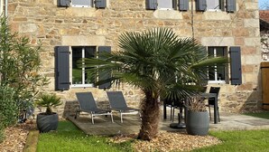 A stone building with a sloped roof, multiple windows with shutters, a small garden with a palm tree, and outdoor seating. - Renovated house, seaside on the port of Ploumanac'h - Perros-Guirec (Perros-Guirec)