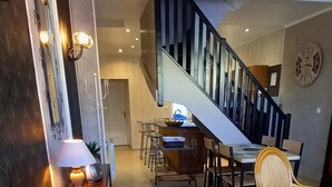 Een ruime woonkamer met een trap, een eettafel met stoelen, een rieten bankje en een klok aan de muur.