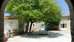 An arched stone structure framing a courtyard with a large tree, a bench, and a building with a tiled roof. - Charming furnished apartment in Mas, UzËs, pool, 656 yards away from town centre (Uzès)