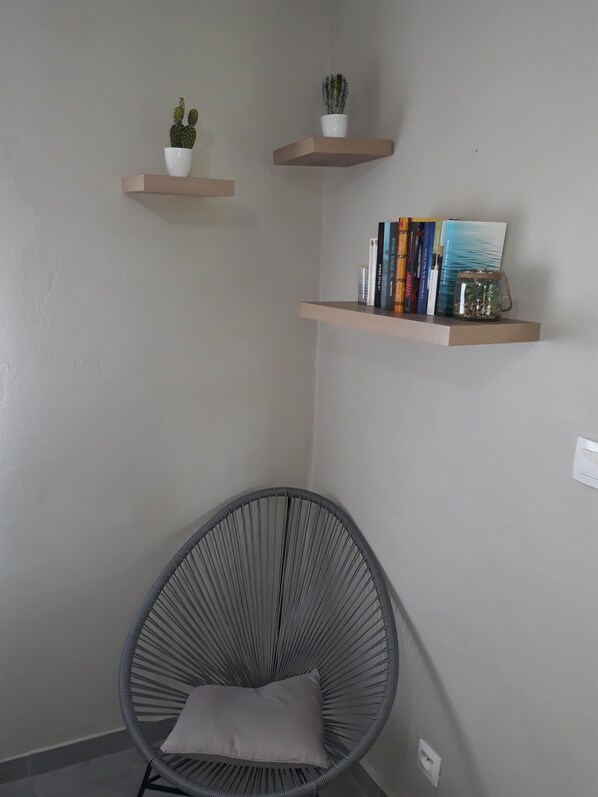 A corner room with a grey woven chair, a cushion, two wall-mounted shelves with plants and books, and a power outlet. - NEW SUITE COTTAGE F3 ANY COMFORT NEAR BEACH ANSE FIGUIER (Rivière-Pilote)