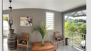 A balcony with wooden chairs, a wooden table, a potted plant, and a painting on the wall. - NEW SUITE COTTAGE F3 ANY COMFORT NEAR BEACH ANSE FIGUIER  (Rivière-Pilote)