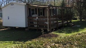 A small cabin with a porch and a fenced deck, surrounded by trees and grass. - Mobile home with air conditioning for 6 people near EUROPA PARK (Boofzheim)