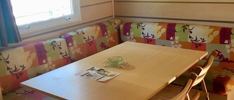 A corner room with a wooden table, a bench with floral upholstery, a window, and a shelf.