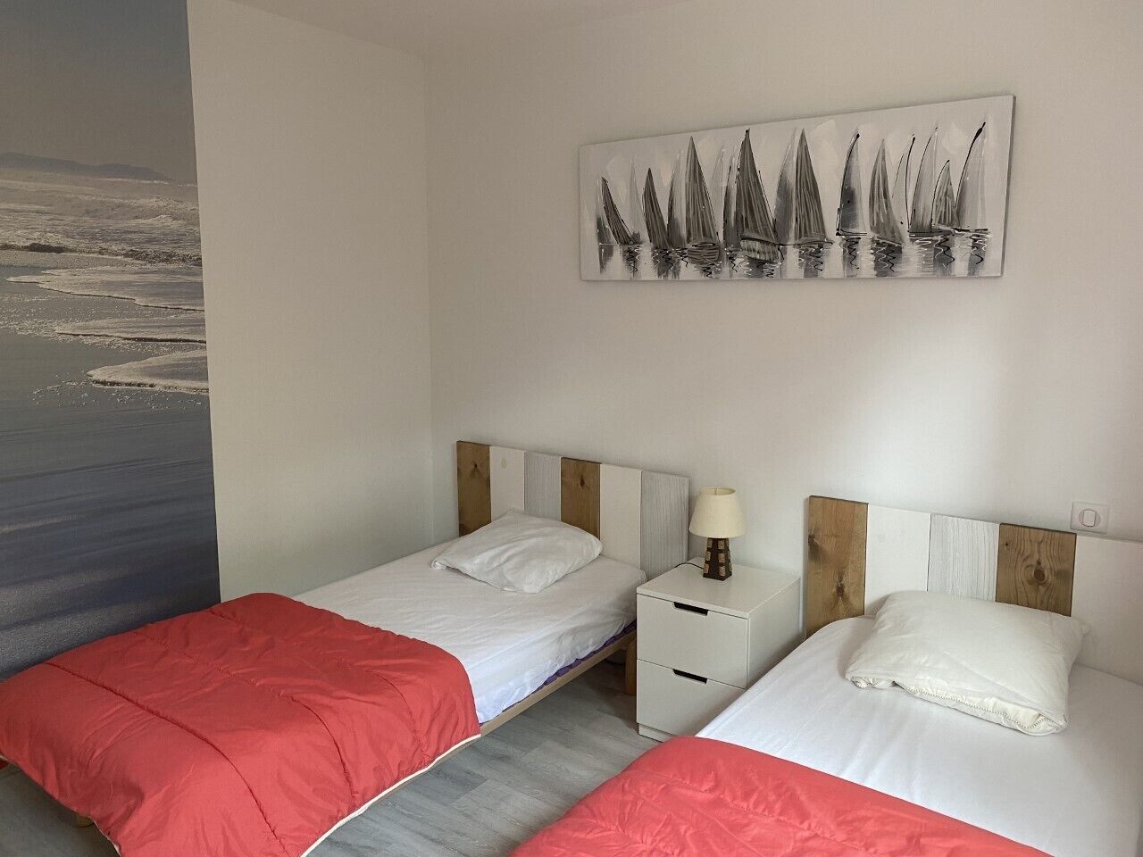 A room with two beds, each with a wooden headboard, white bedding, and a red blanket. There is a nightstand with a lamp and a painting of sailboats on the wall.