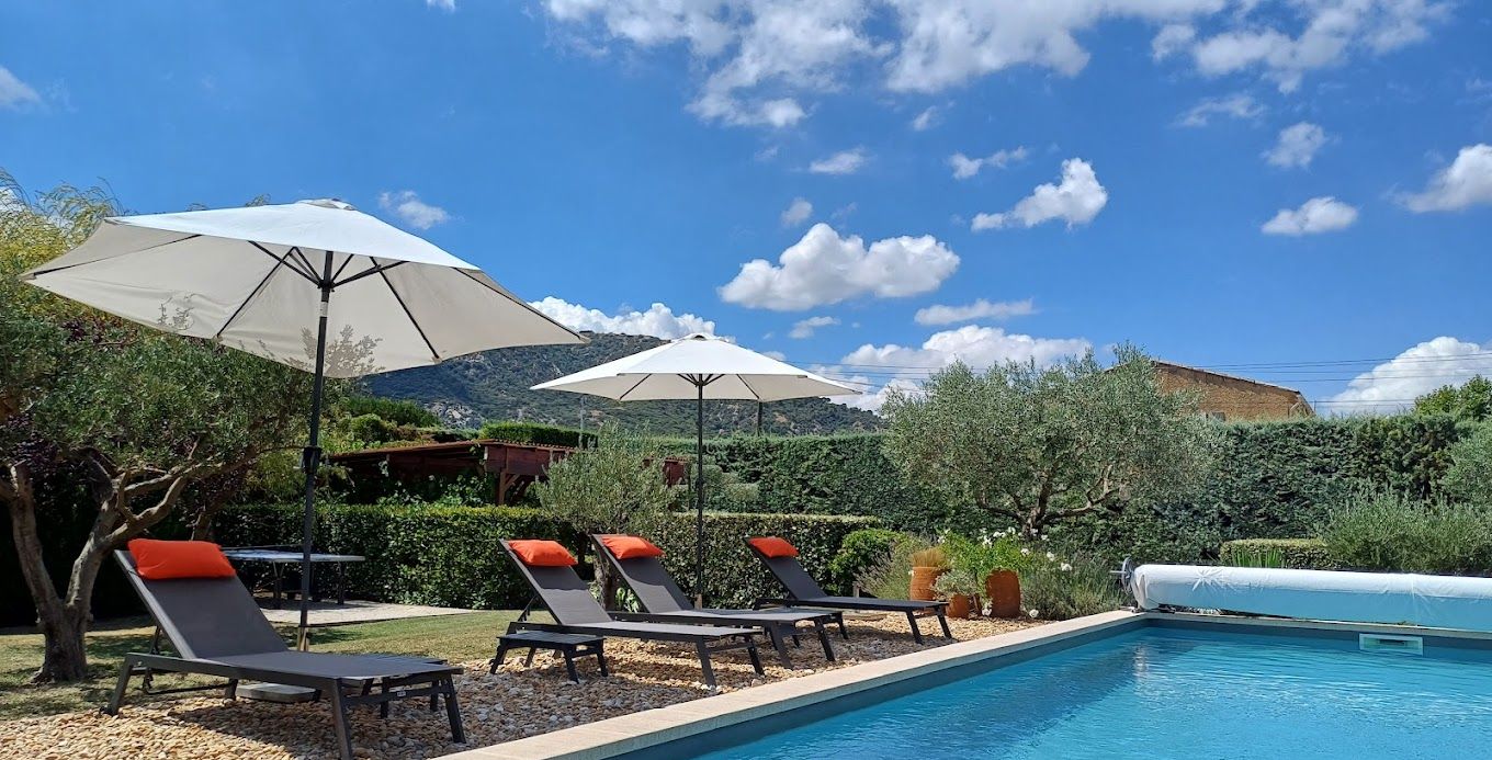 Zona de piscina con sillones de playa y sombrillas, rodeada de vegetación y con montañas al fondo.