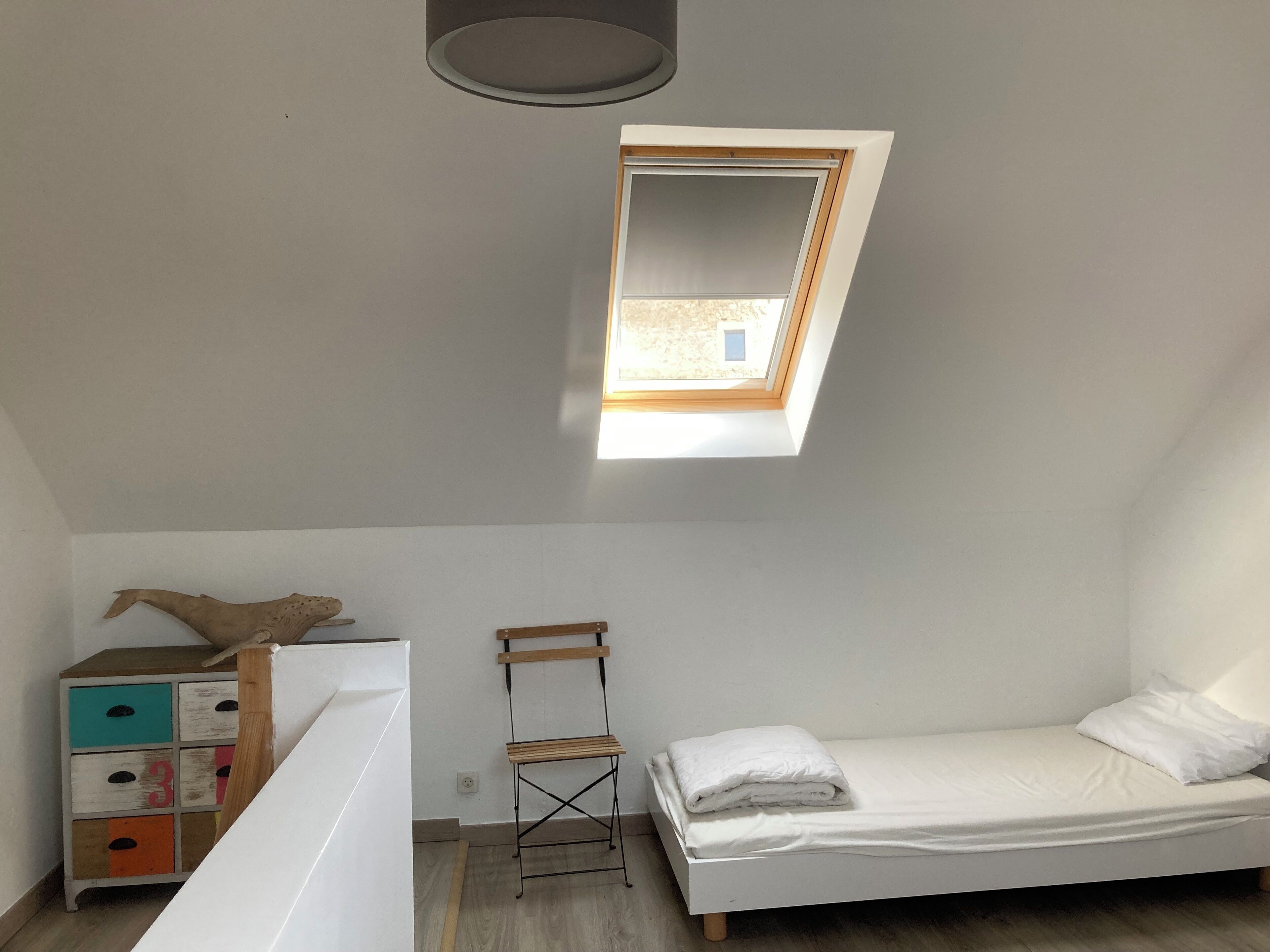 A small, bright room with a skylight, a bed, a wooden chair, and a colorful dresser.