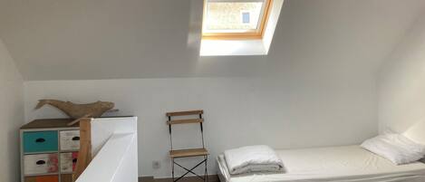 A small, bright room with a skylight, a bed, a wooden chair, and a colorful dresser.