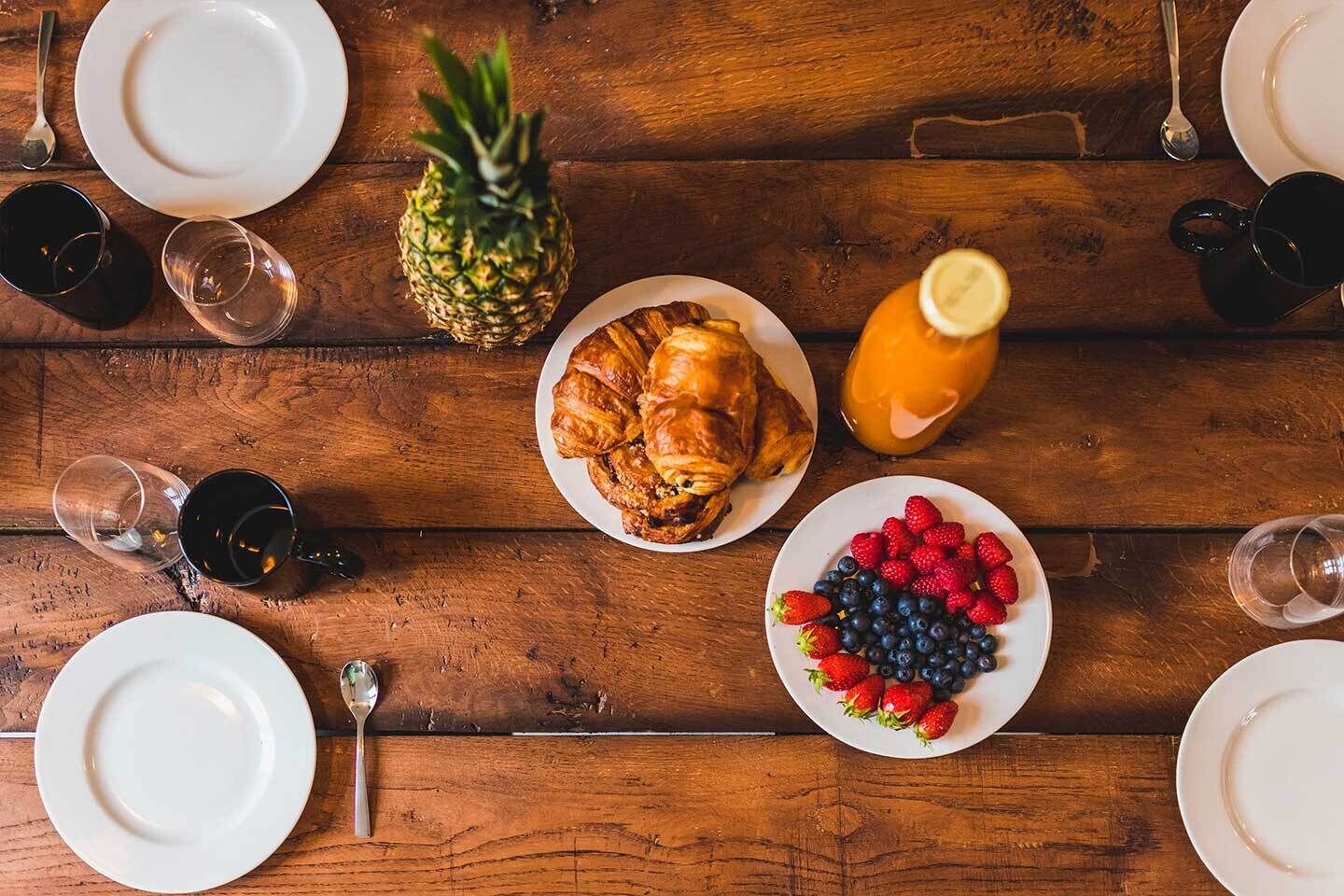 Una mesa de madera con alimentos para el desayuno, incluyendo croissants, una piña y una fuente de bayas.