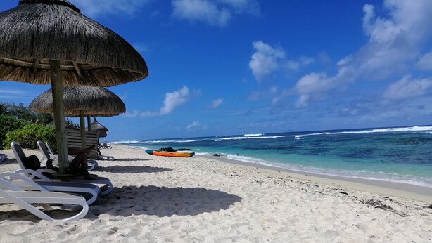 Ein Strand mit weißen Liegestühlen, einem Reetdachschirm und einem Kajak im Sand.