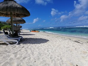 Una playa con sillas de playa blancas, una sombrilla de paja y un kayak en la arena.