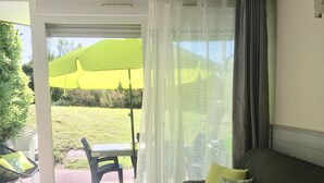 A living room with a dark-colored sofa, a small table with a vase, and a view of a green lawn and outdoor furniture through a glass door. - Appt classified 4 *, 2 swimming pools, free WIFI, terrace, bbq, edge of the marine lake (Vieux-Boucau-Les-Bains)