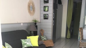 A living room with a dark sofa, two small tables with potted plants, a wicker chair, a green curtain, and a refrigerator. - Appt classified 4 *, 2 swimming pools, free WIFI, terrace, bbq, edge of the marine lake (Vieux-Boucau-Les-Bains)
