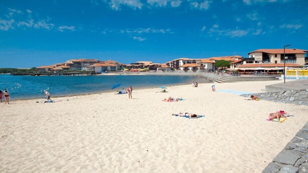 A beach with people sunbathing and swimming, buildings with red roofs, and a clear blue sky. - Appt classified 4 *, 2 swimming pools, free WIFI, terrace, bbq, edge of the marine lake (Vieux-Boucau-Les-Bains)