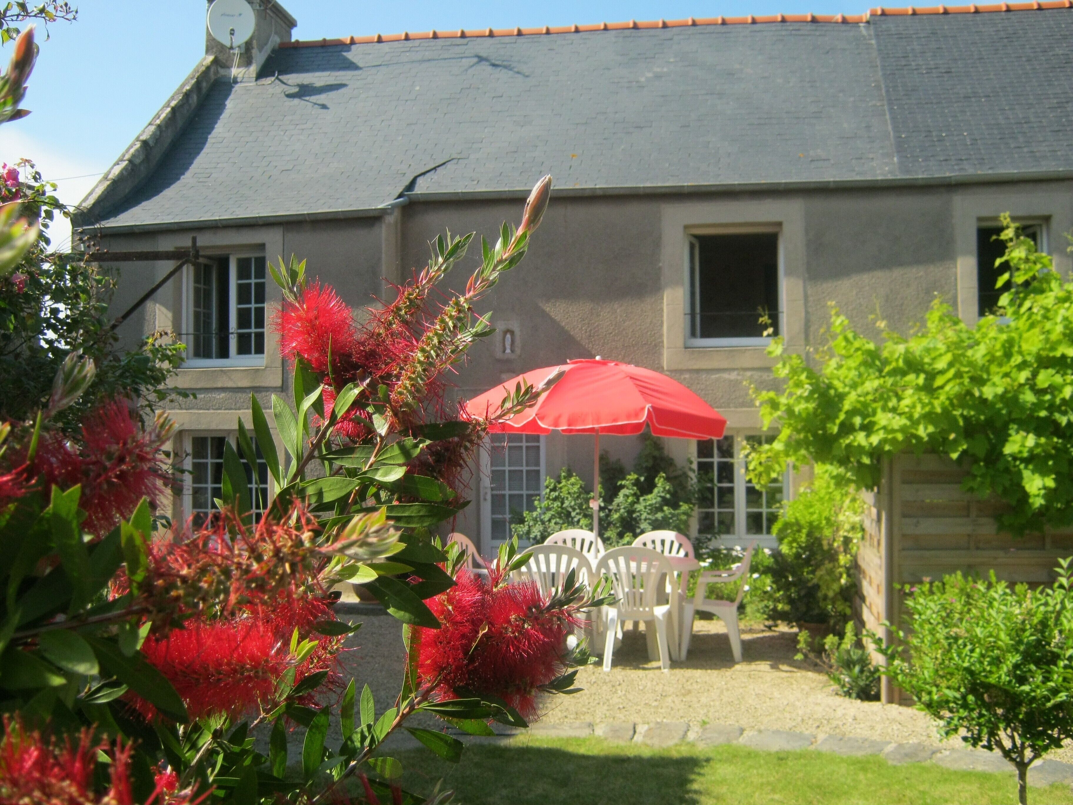 Une maison avec un toit gris, un parapluie rouge et des chaises blanches dans le jardin.