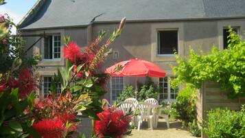 Une maison avec un toit gris, un parapluie rouge et des chaises blanches dans le jardin.