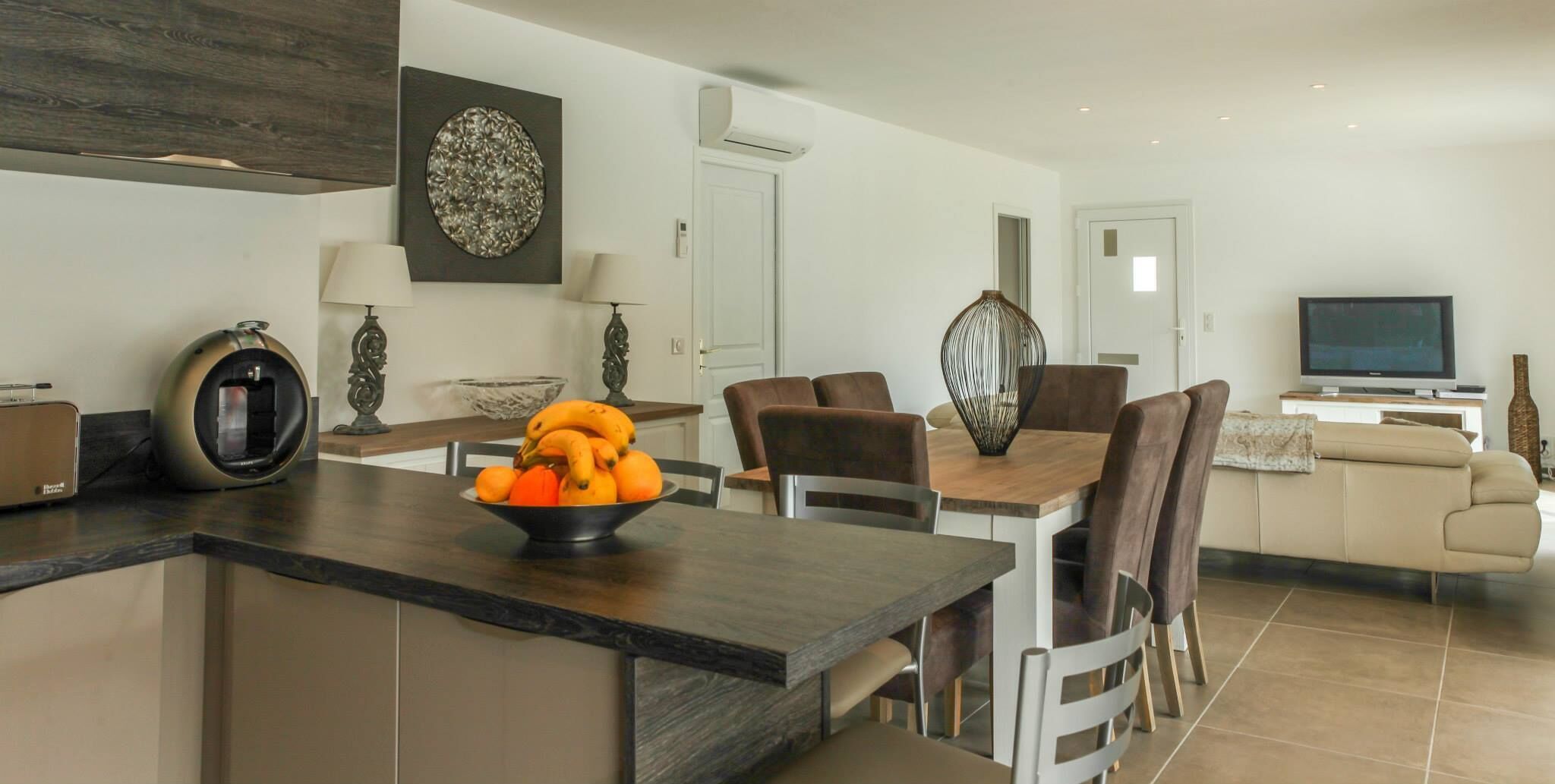 A modern kitchen with a central island, dining area with chairs, a sofa, and a TV in the living room.
