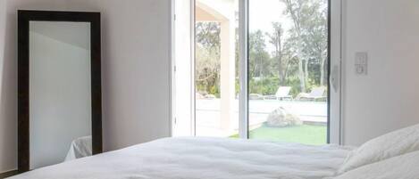 A bedroom with a large bed, a sliding glass door leading to an outdoor area with a pool and greenery.