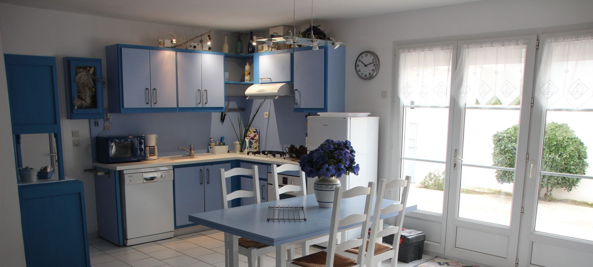 Une cuisine de taille réduite avec des meubles bleus, une table à manger et des chaises. Il y a une horloge au mur et une fenêtre avec des rideaux.